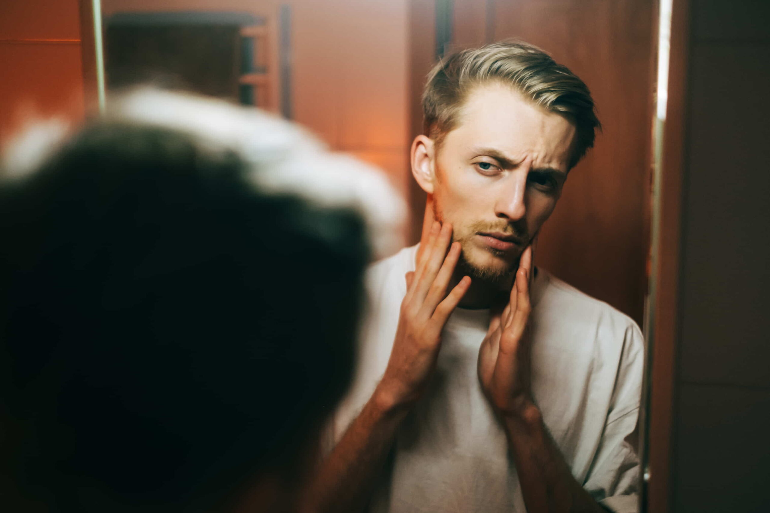 Text: A man standing in front of the mirror, touching his face and analyzing its shape.
